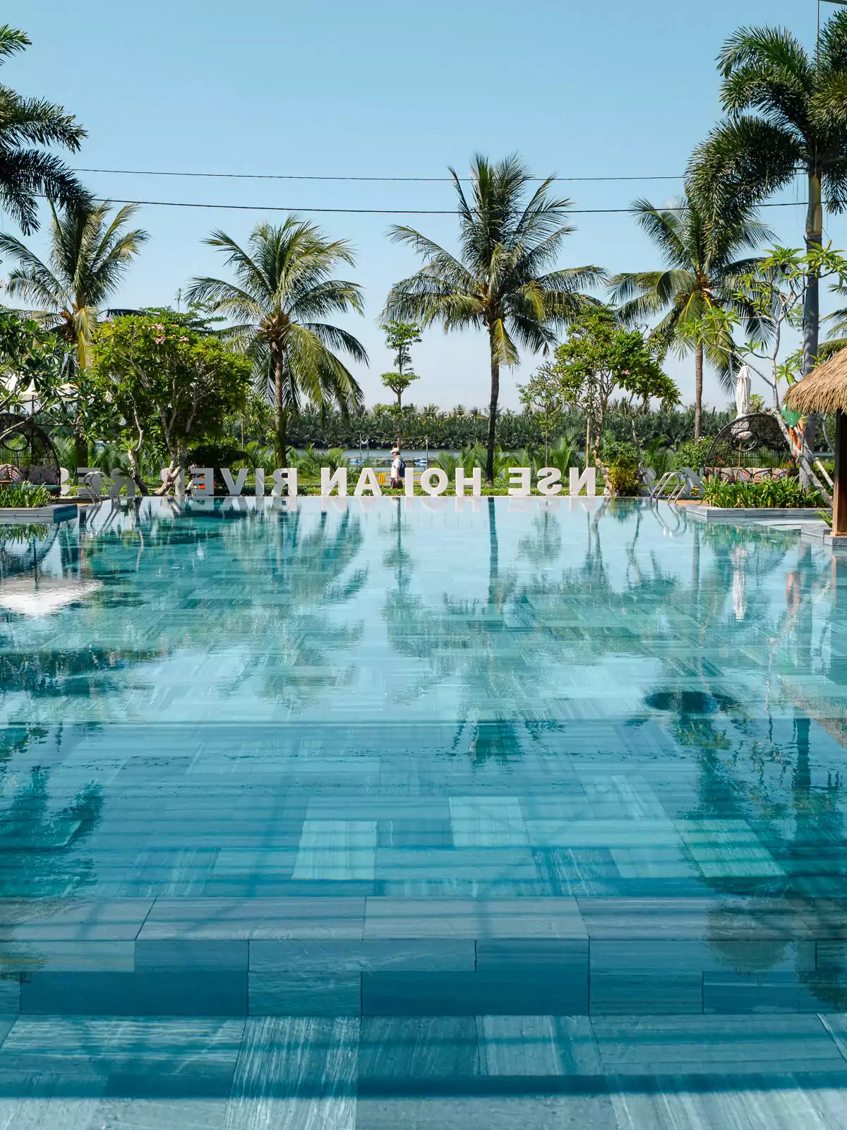 A luxurious pool at a resort in Hoi An, Vietnam, lined with palm trees and reflecting the clear blue sky, ideal for a relaxing getaway.