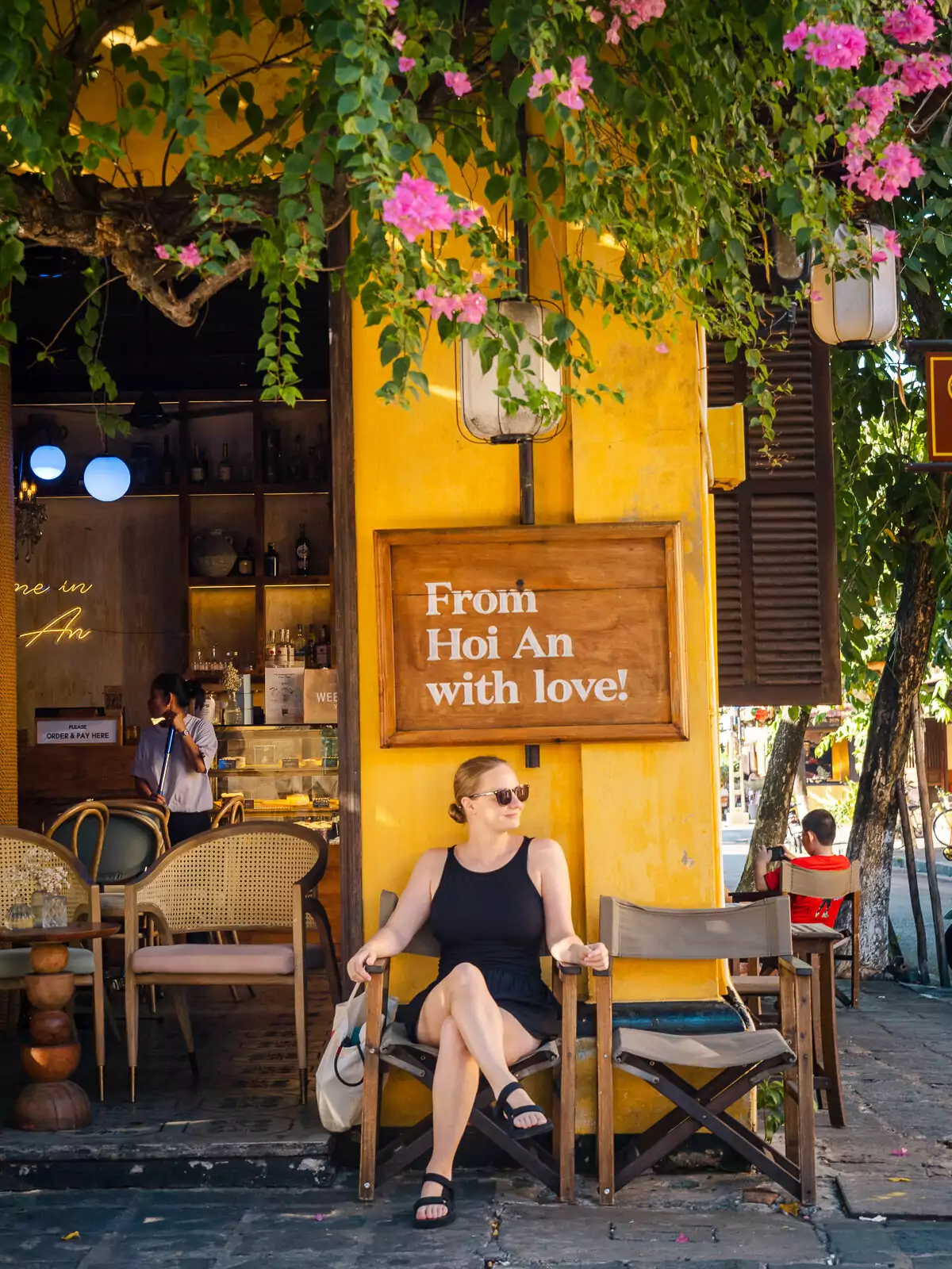 A woman relaxing in front of a vibrant yellow café in Hoi An with a sign that reads "From Hoi An with Love," capturing the charm of the city.