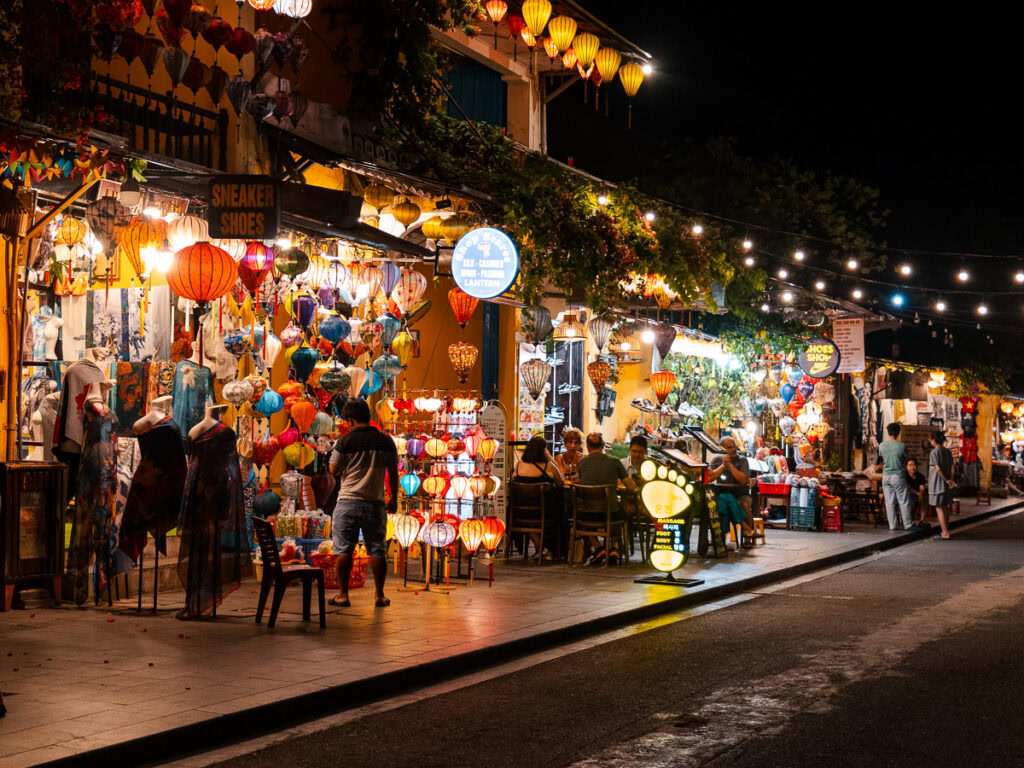 The vibrant lantern-lit shops of Hoi An at night, where visitors browse colourful souvenirs and enjoy the lively evening atmosphere.