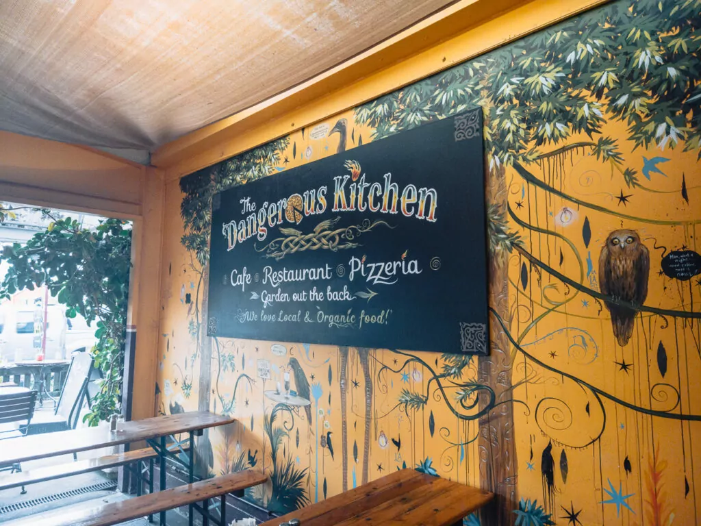 Colourful mural wall and sign at The Dangerous Kitchen café in Golden Bay, with vibrant art, fairy lights, and wooden tables in the cosy dining space