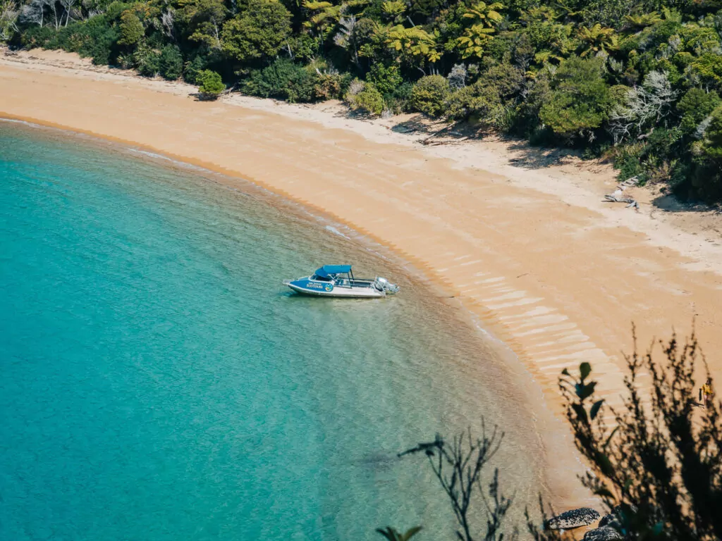 A small boat is anchored off a crescent-shaped golden beach surrounded by lush native forest in Abel Tasman National Park