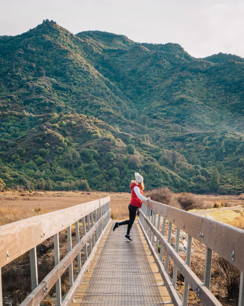 A woman in a red vest pauses on a footbridge with lush green hills behind her near the start of the Abel Tasman Coast Track in Marahau