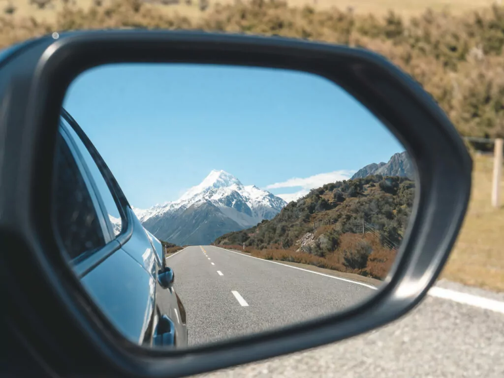 Snow-capped Aoraki Mount Cook reflected in a car's side mirror while driving along the highway through the Mackenzie Basin