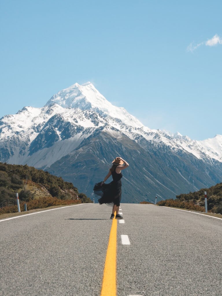 Alexx standing on the road to Aoraki Mount Cook with the snow-capped peak towering in the distance