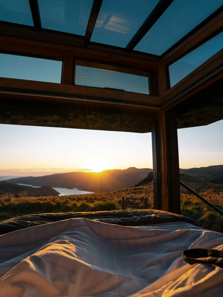 View from a glass cabin bed overlooking a glowing sunrise above hills and Port Levy harbour on the Banks Peninsula near Christchurch