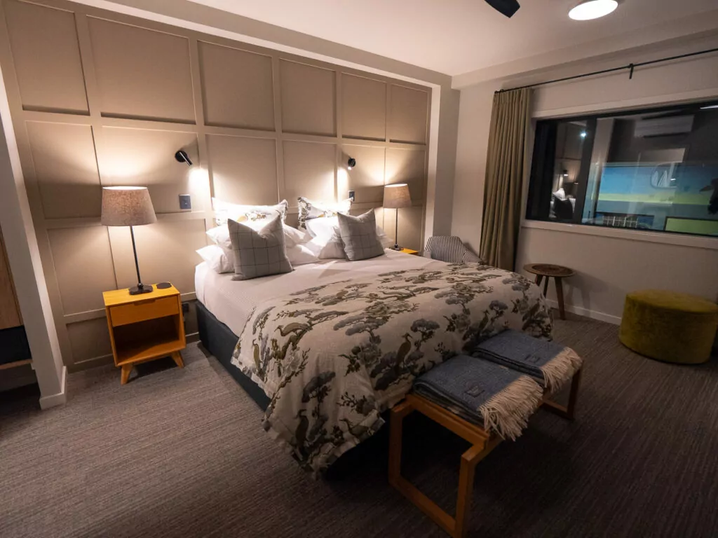 A warmly lit hotel room in Blenheim featuring a large bed with botanical bedding, soft lighting, and a modern window view.