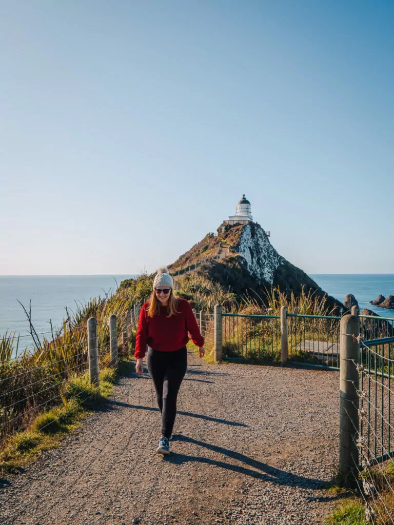 Woman in red jacket walking along gravel path towards the iconic Nugget Point Lighthouse on a narrow peninsula with ocean on both sides