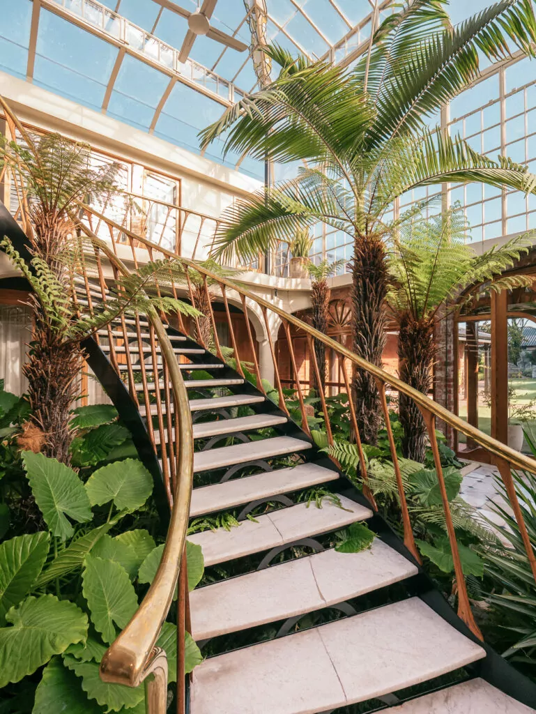 A grand curved staircase with golden railings winds through a sunlit conservatory filled with tropical plants and palm trees at Britten Stables in Christchurch