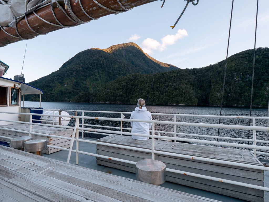 A woman sits wrapped in a blanket on the deck of a boat, looking out over the calm, remote waters of Doubtful Sound at sunset