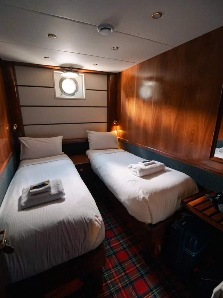 A cozy twin cabin with wooden walls, tartan carpet, and a small porthole aboard a Doubtful Sound overnight cruise ship