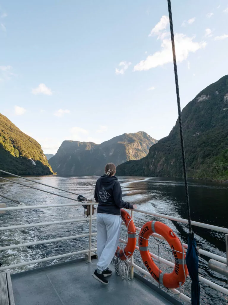 Woman in dark hoodie standing on boat deck facing towering forested mountains that rise vertically from Doubtful Sound's calm waters in Fiordland