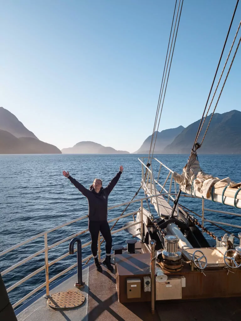 A woman stands with arms raised on the bow of a cruise boat in Doubtful Sound in Fiordland National Park, surrounded by morning light and dramatic fjord scenery