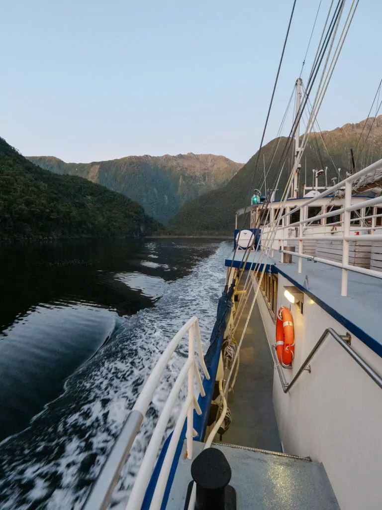 A cruise boat sails through the narrow passageways of Doubtful Sound, flanked by towering cliffs and glowing evening light