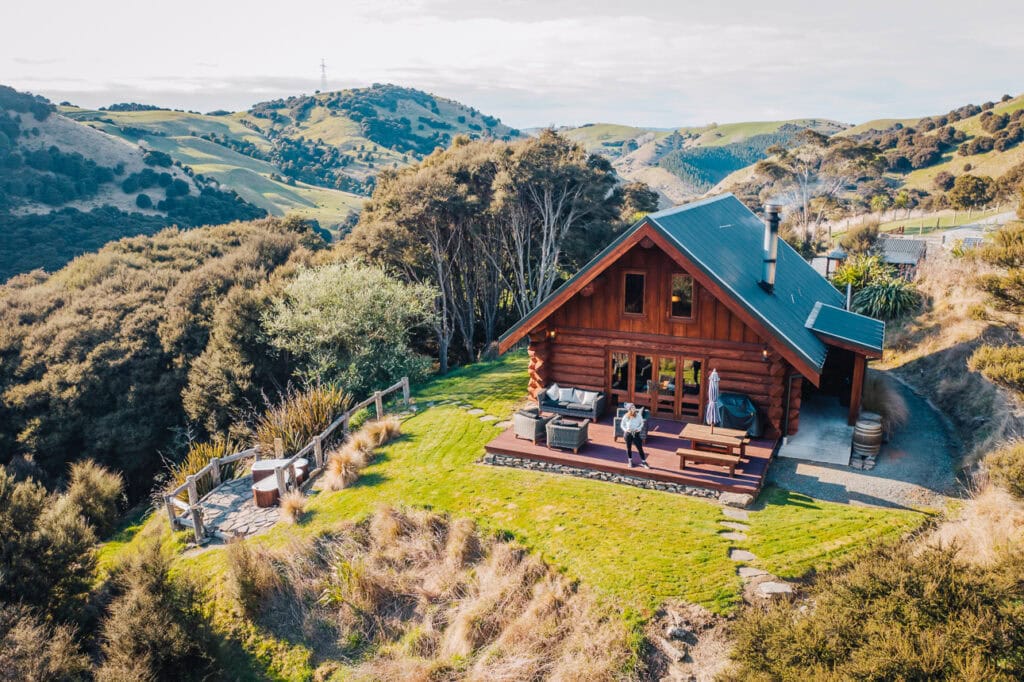 Aerial view of a rustic wooden lodge Cascade Creek near Dunedin, nestled in rolling green hills with sheep grazing and dramatic coastal landscape in the background