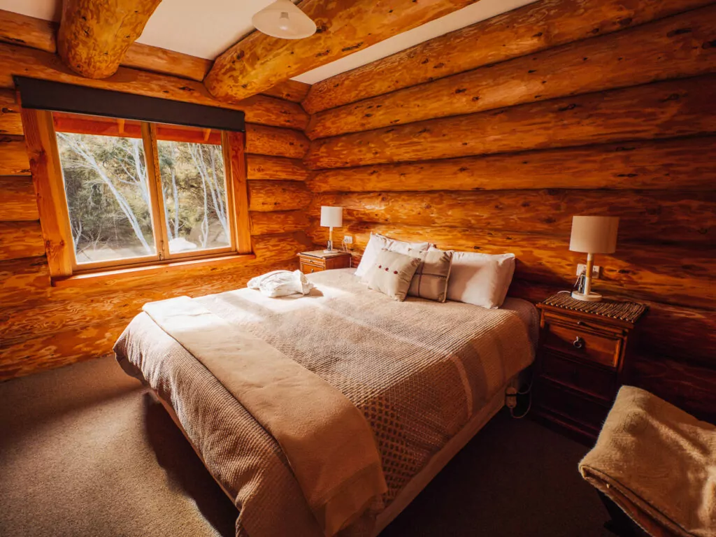 Cosy log cabin bedroom with exposed wooden beams, white bedding on a queen bed, and large windows overlooking mountains