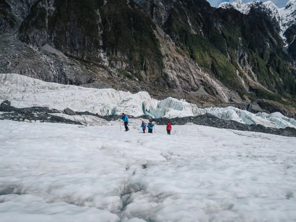 A small group of hikers trek across the icy surface of Franz Josef Glacier, with jagged ice formations and steep cliffs rising around them
