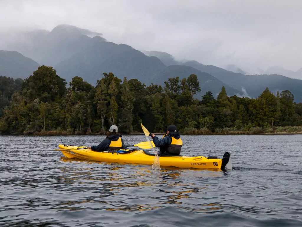 Two people paddle a bright yellow kayak on a still lake surrounded by forest and misty mountains near Franz Josef