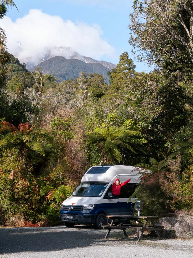 A campervan parked beside native forest at a holiday park near Franz Josef, with a person in red waving and mountain views in the background