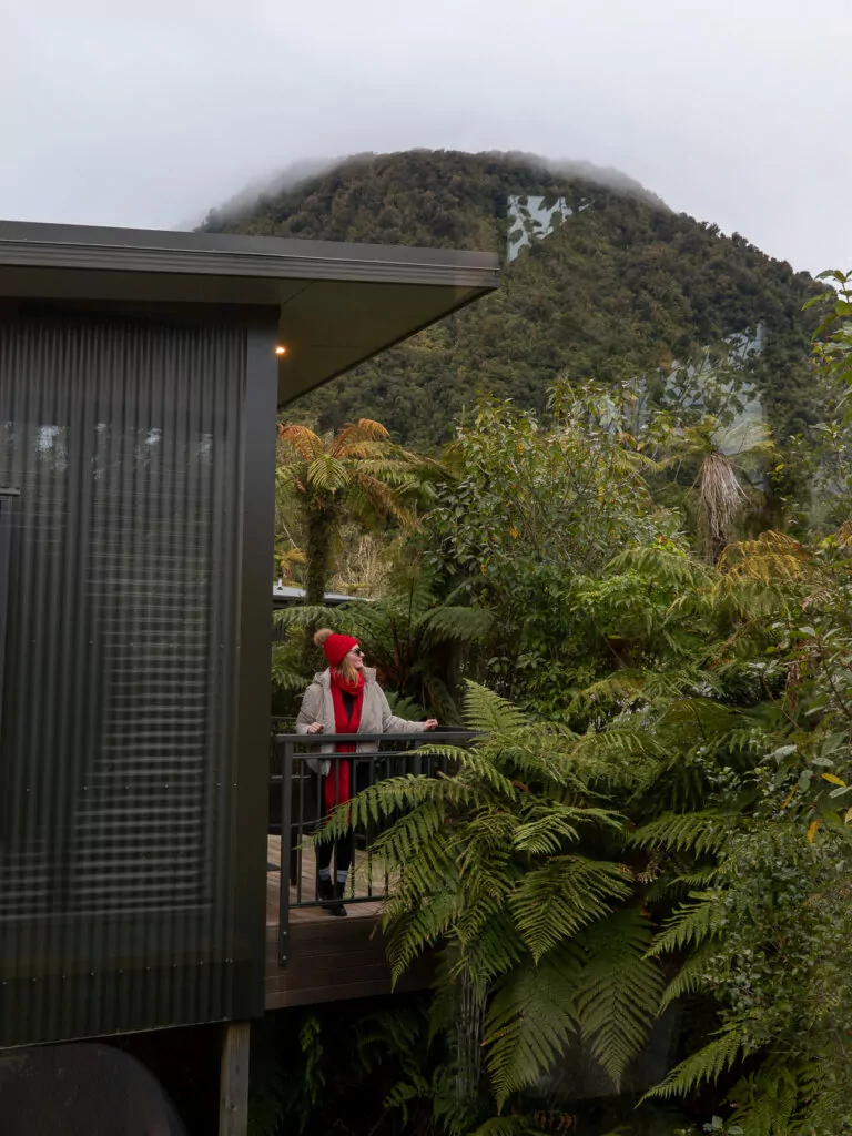 A woman stands on a private balcony at Rainforest Retreat in Franz Josef, surrounded by lush ferns and native bush, with a misty mountain rising in the background.
