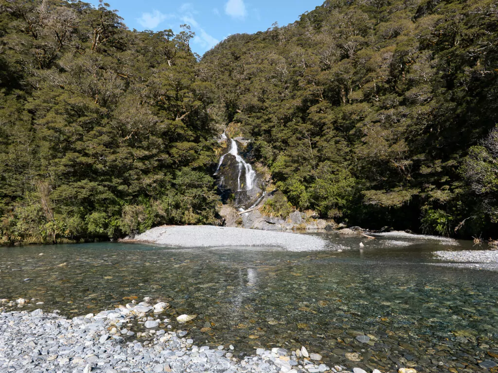 Fantail Falls cascades down a rocky cliff into a clear, shallow river surrounded by dense native forest in Mount Aspiring National Park