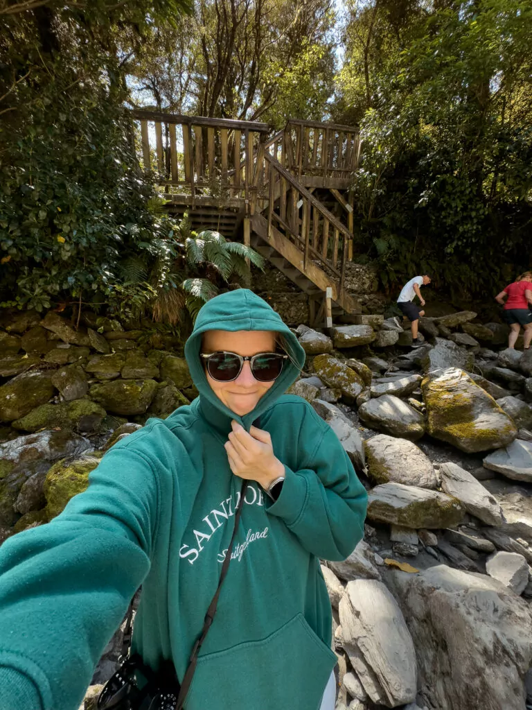 A woman in a green hoodie takes a selfie near the rocky trail and wooden staircase grimacing due to swarms of sandflies at a waterfall along the Haast Pass