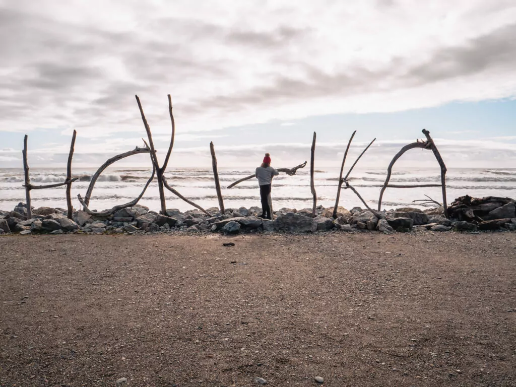 A large driftwood sculpture spelling out "Hokitika" stands on the beach, with a person posing in the middle and the ocean stretching behind