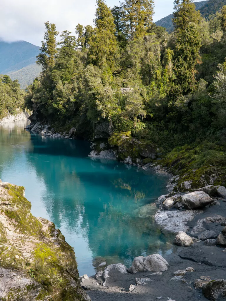 Vibrant turquoise water at the Hokitika Gorge, framed by mossy rocks and dense native bush, under soft mountain light