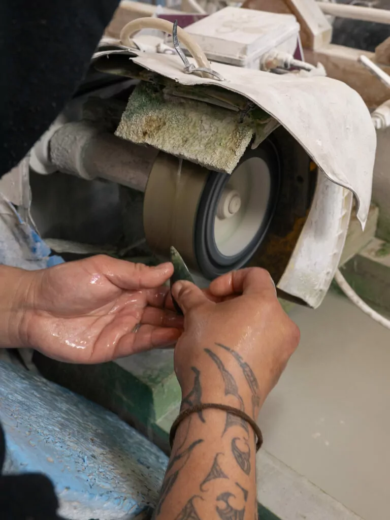 Close-up of a tattooed artist’s hands shaping a piece of pounamu (greenstone) on a grinding wheel in Hokitika, a town known for Māori carving and jade work