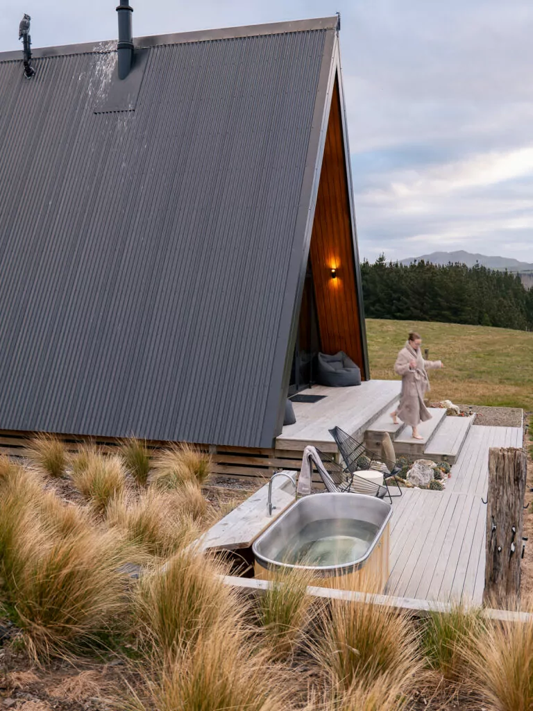 A cozy A-frame cabin set in Hurunui's high country at Tawanui Farm, with a person in a robe walking past a steaming outdoor hot tub on a wooden deck surrounded by tussock grass