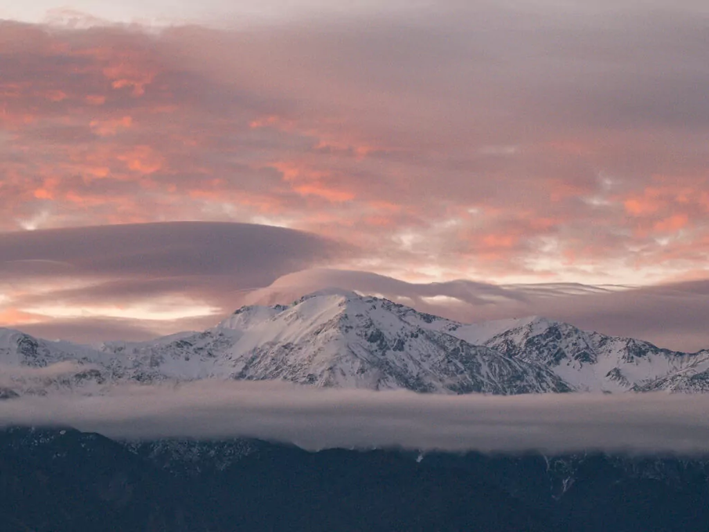 The Seaward Kaikōura Ranges covered in snow beneath a dramatic pink and orange sunset sky, with low clouds drifting along the foothills