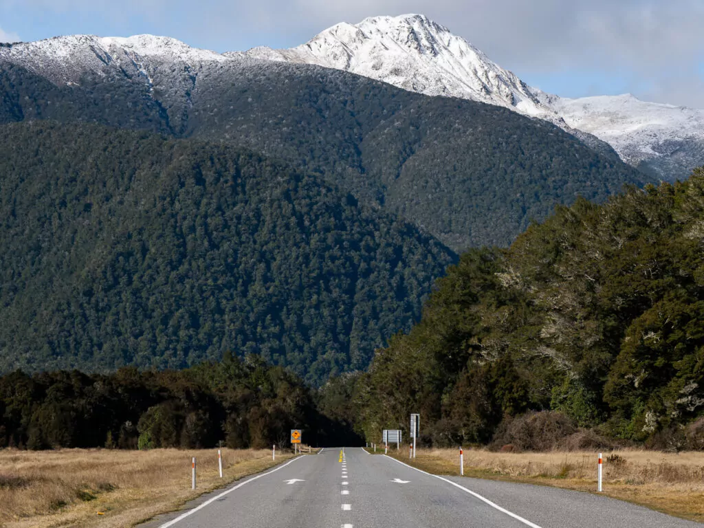 A straight mountain highway cuts through Lewis Pass, with snow-capped peaks and dense forest rising on either side under a bright sky.