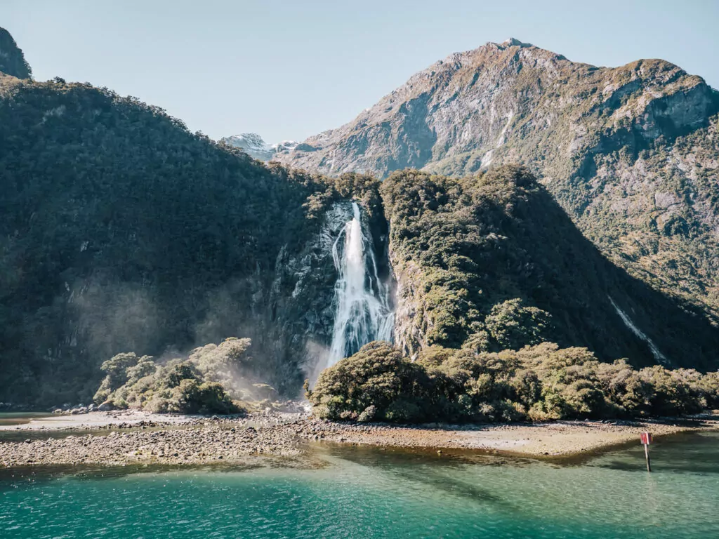 Stirling Falls plunging into the turquoise waters of Milford Sound with rocky mountain peaks and native forest in Fiordland National Park
