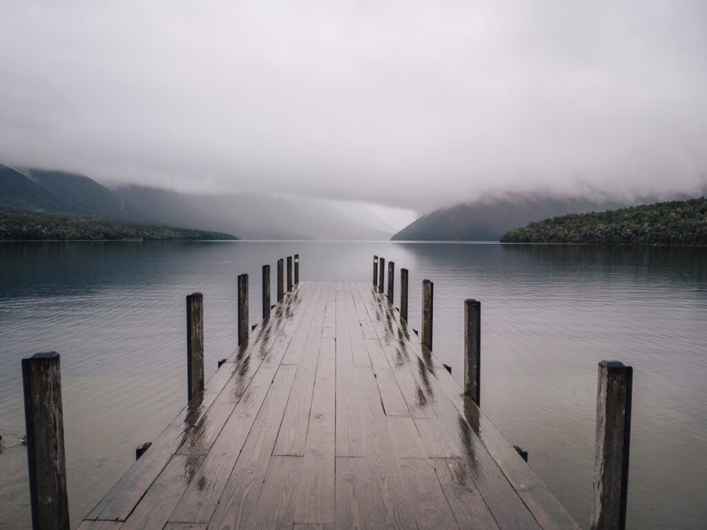 A wooden jetty stretches into the still, mist-covered waters of Lake Rotoiti in Nelson Lakes National Park, framed by forested mountains