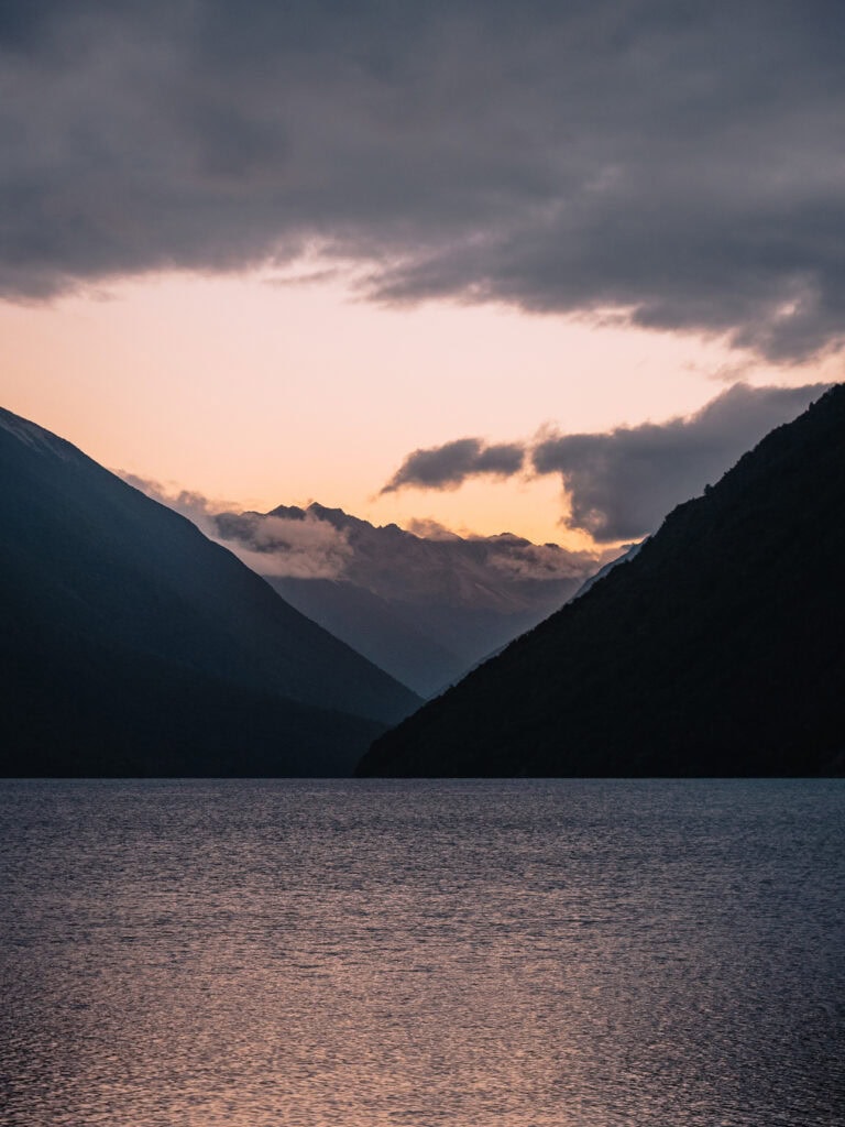 Soft sunset light filters through a mountain pass at Lake Rotoiti, with dark silhouettes of the surrounding peaks reflected in the water