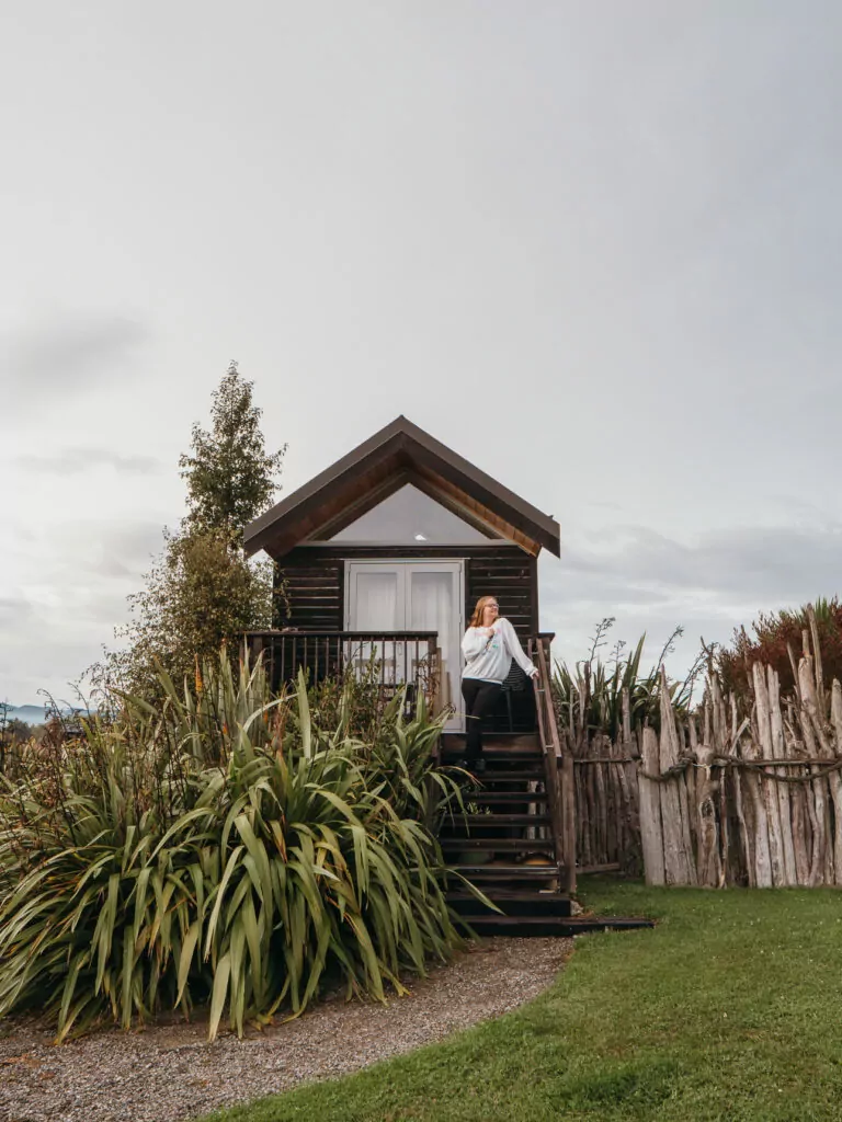 A woman sits on the steps of a rustic wooden cabin at Rabbit Island Huts near Nelson, surrounded by native flax plants and a wooden fence under a cloudy sky