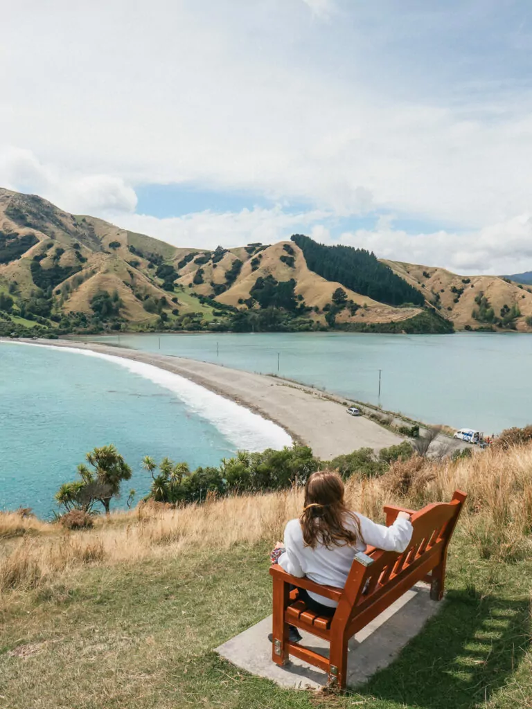 A woman sits on a wooden bench looking out over the turquoise waters and curved sandbar of Cable Bay near Nelson, framed by golden coastal hills