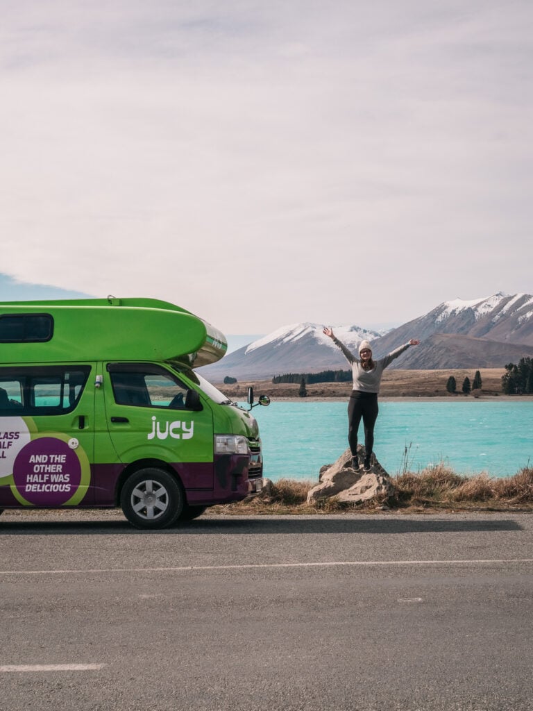 Alexx standing with arms raised beside a green Jucy campervan parked in front of Lake Tekapo