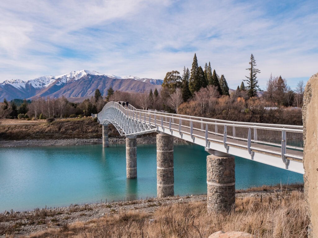 Footbridge crossing the Tekapo River towards the Church of the Good Shepherd with snow-capped mountains behind