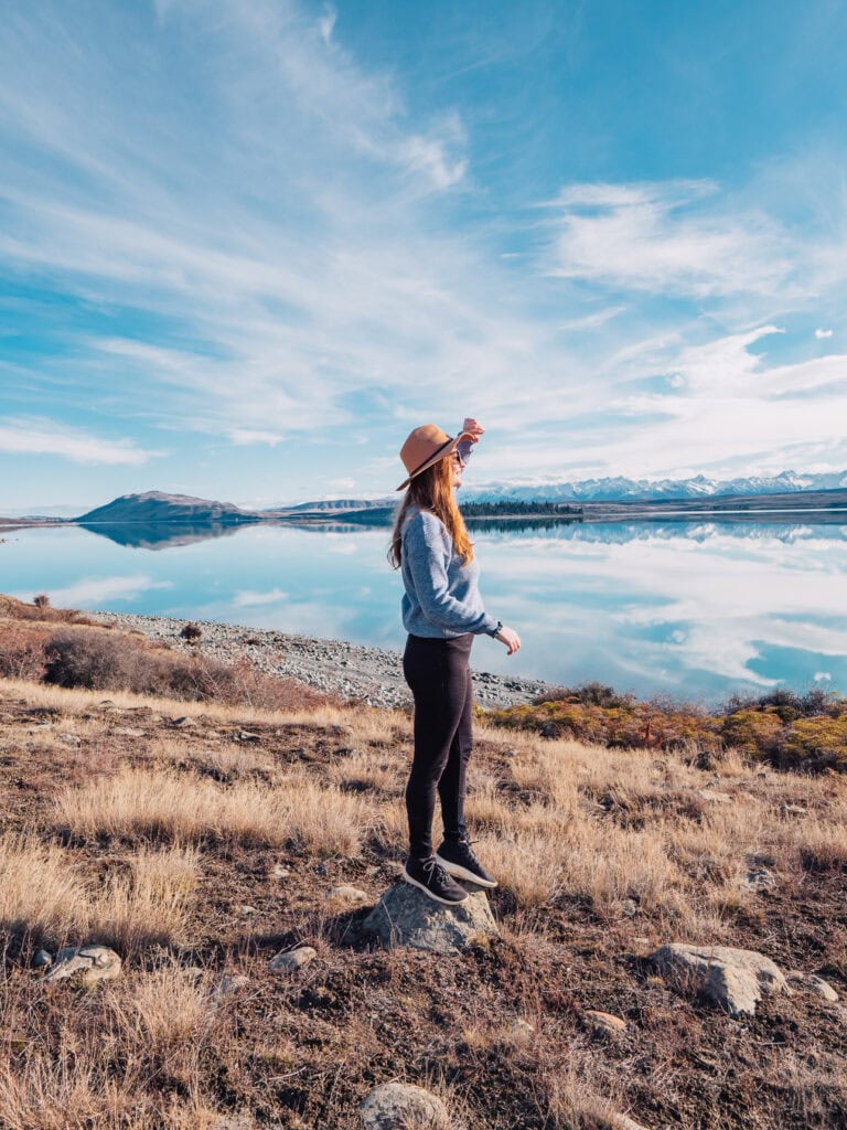 Alexx standing on a rock beside Lake Tekapo with mountains reflected in the glassy water
