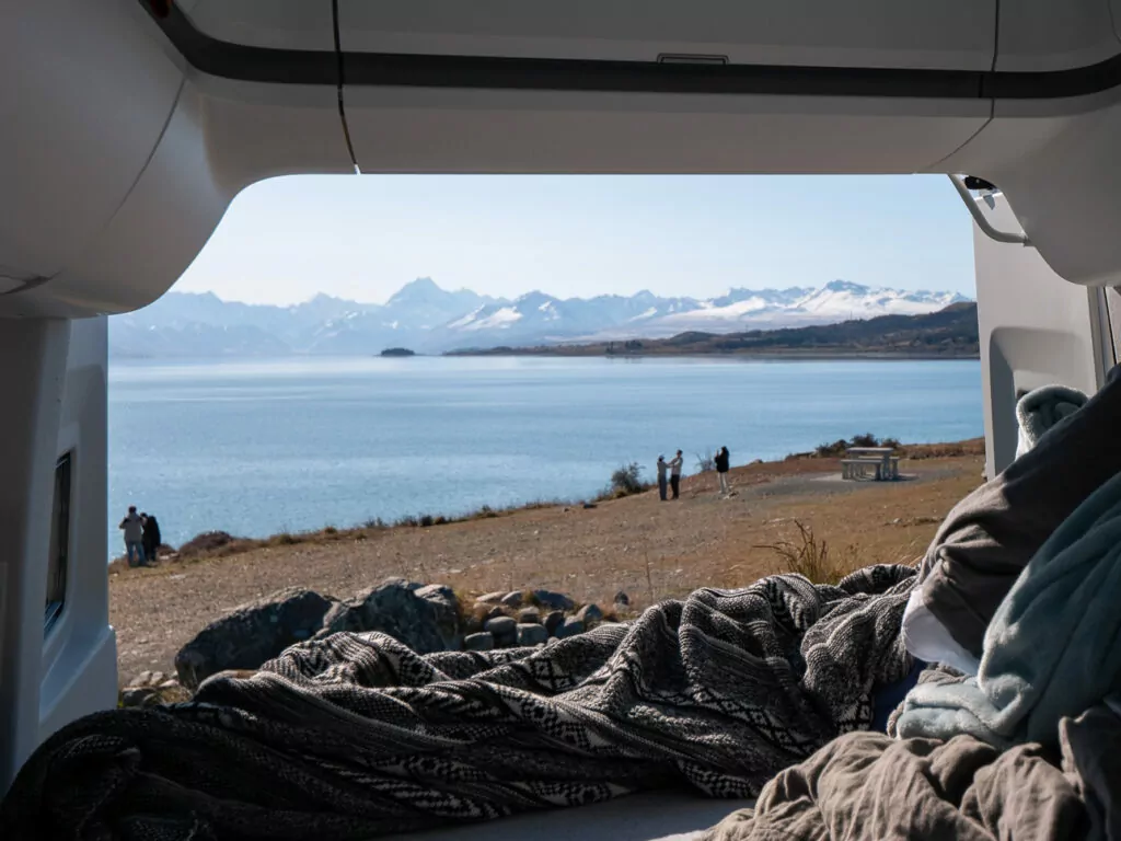 View from inside a campervan with rumpled bedding in foreground looking out to the brilliant turquoise waters of Lake Pukaki with people on the pebbly shore