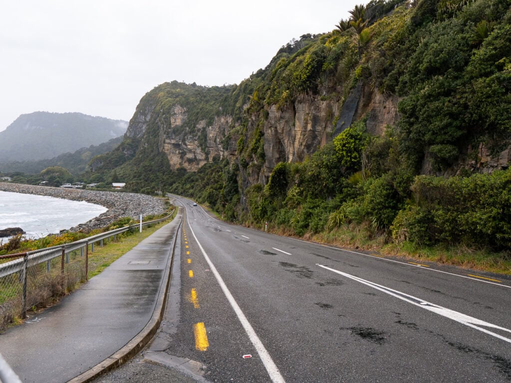 A winding road hugs the cliffs above the ocean near Punakaiki, with lush vegetation and misty hills lining the dramatic West Coast drive