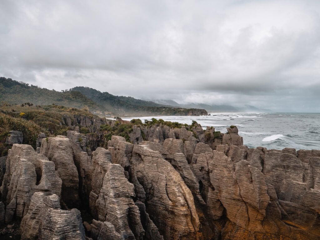 The layered limestone formations of the Pancake Rocks rise from the West Coast shoreline at Punakaiki, with waves crashing below under grey skies