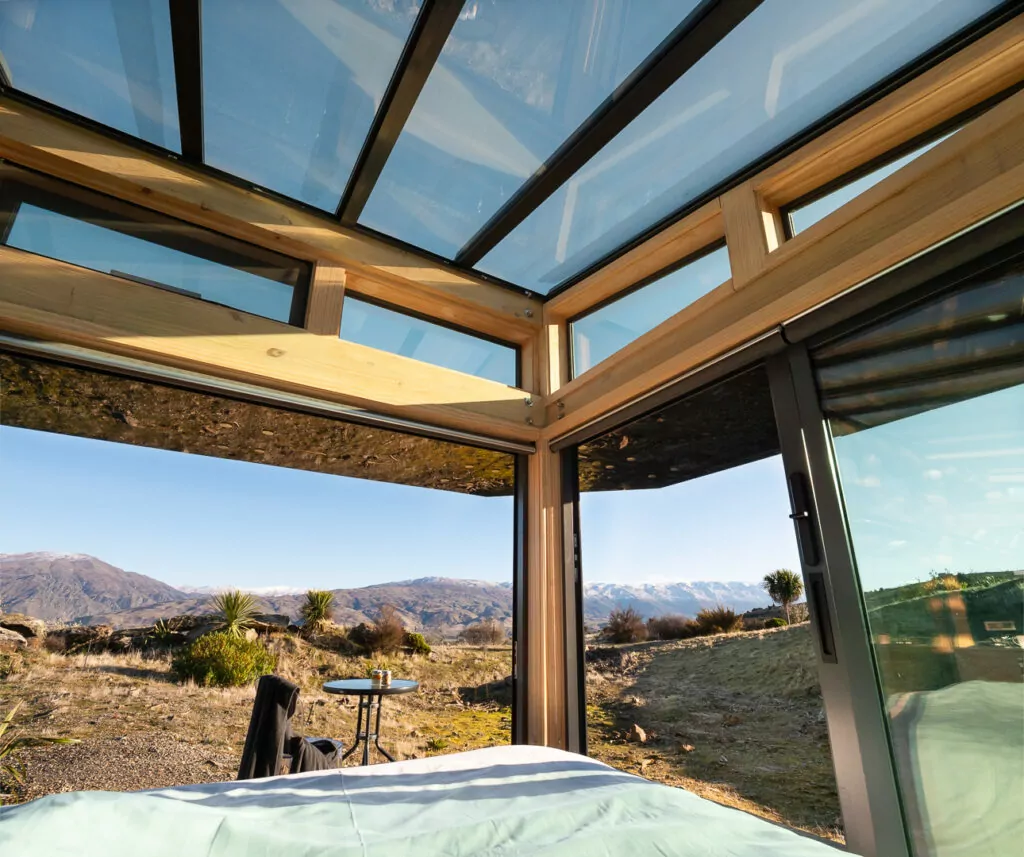 Modern glass-walled eco cabin with floor-to-ceiling windows and outdoor deck furniture overlooking golden tussock hills and distant mountains at Taima PurePod near Cromwell