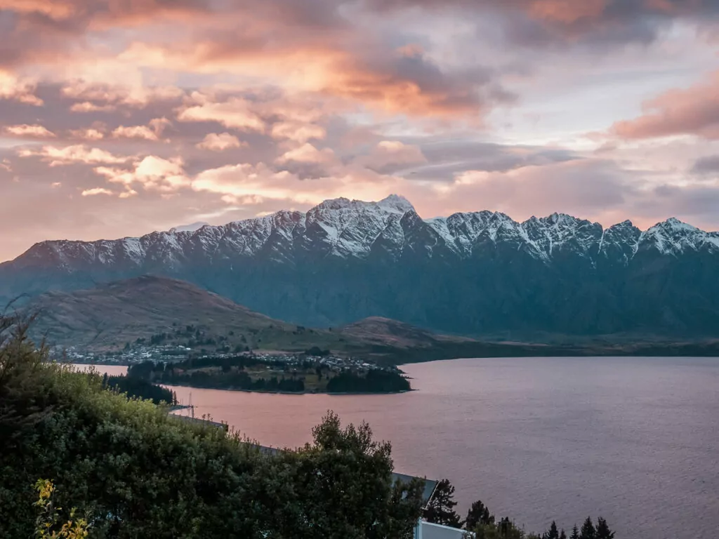 Panoramic sunset view over Queenstown and Lake Wakatipu with snow-capped mountains.
