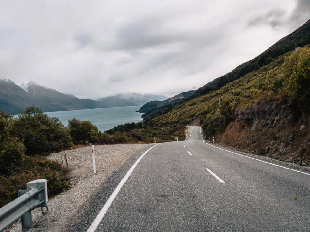 A winding road leads toward Lake Wakatipu with mountain views and cloudy skies near Glenorchy, part of one of the South Island’s most scenic drives