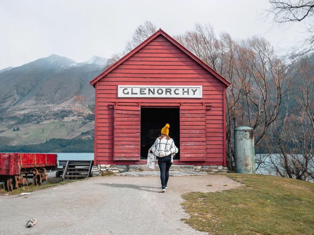 A woman in a yellow beanie walks toward the historic red Glenorchy Shed, set against snow-dusted mountains on the edge of Lake Wakatipu