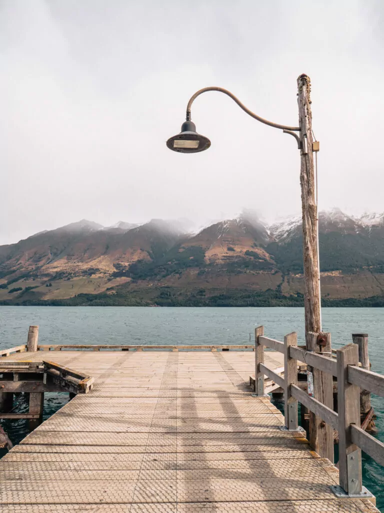 A vintage-style lamp stands on the wooden jetty at Glenorchy Wharf, overlooking the lake with misty mountains in the background