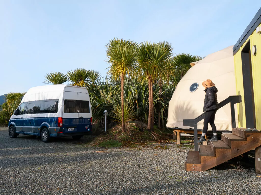 A woman steps down from a glamping dome at Ross Beach TOP 10 Holiday Park, with a campervan parked nearby among cabbage trees and gravel paths