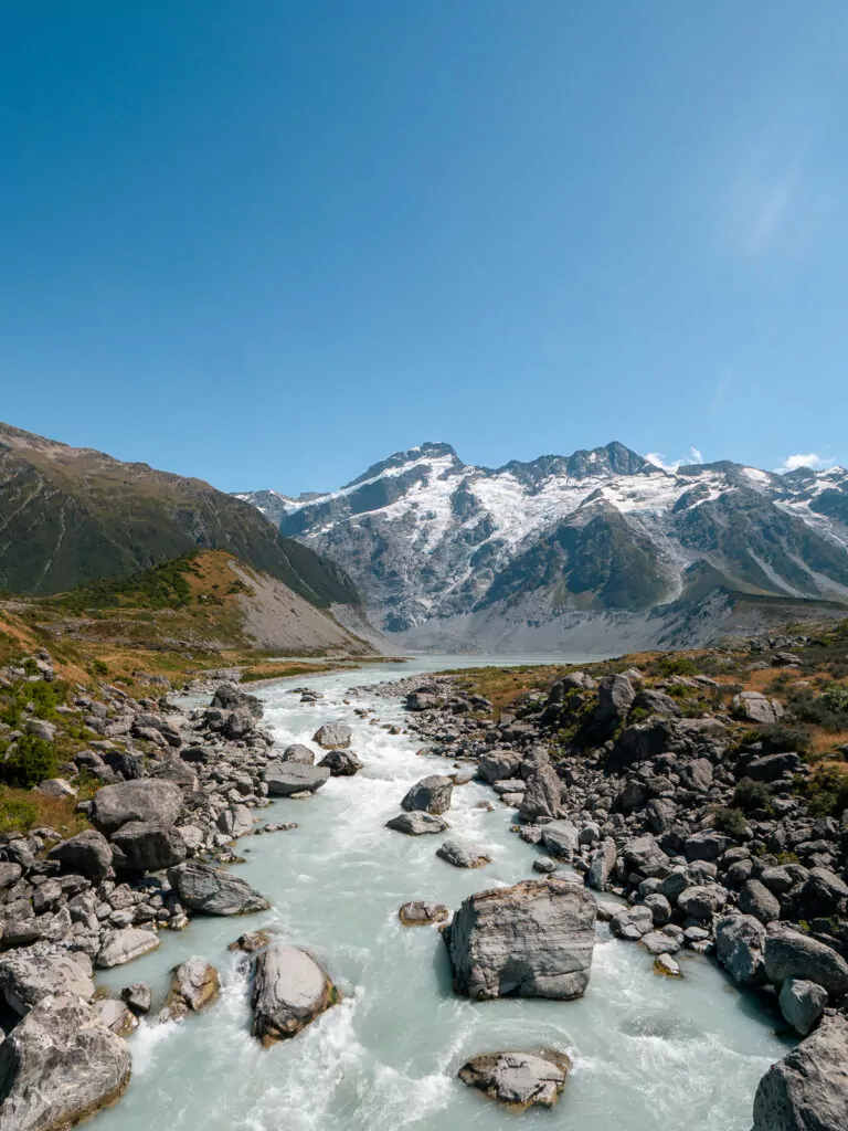 A glacial river winds through rocky terrain toward the snow-covered peaks of Aoraki / Mount Cook National Park, viewed from the Hooker Valley Track on a sunny day
