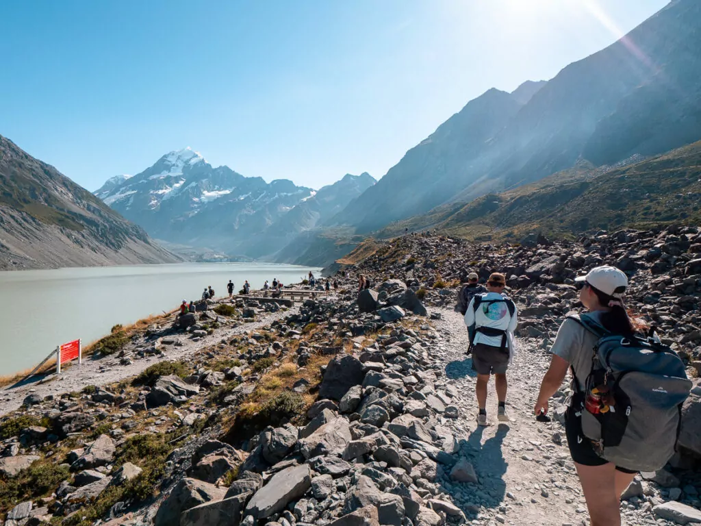 Hikers walking along the rocky shore of Hooker Lake with icebergs floating in the glacial water and Aoraki Mount Cook rising behind in Mount Cook National Park